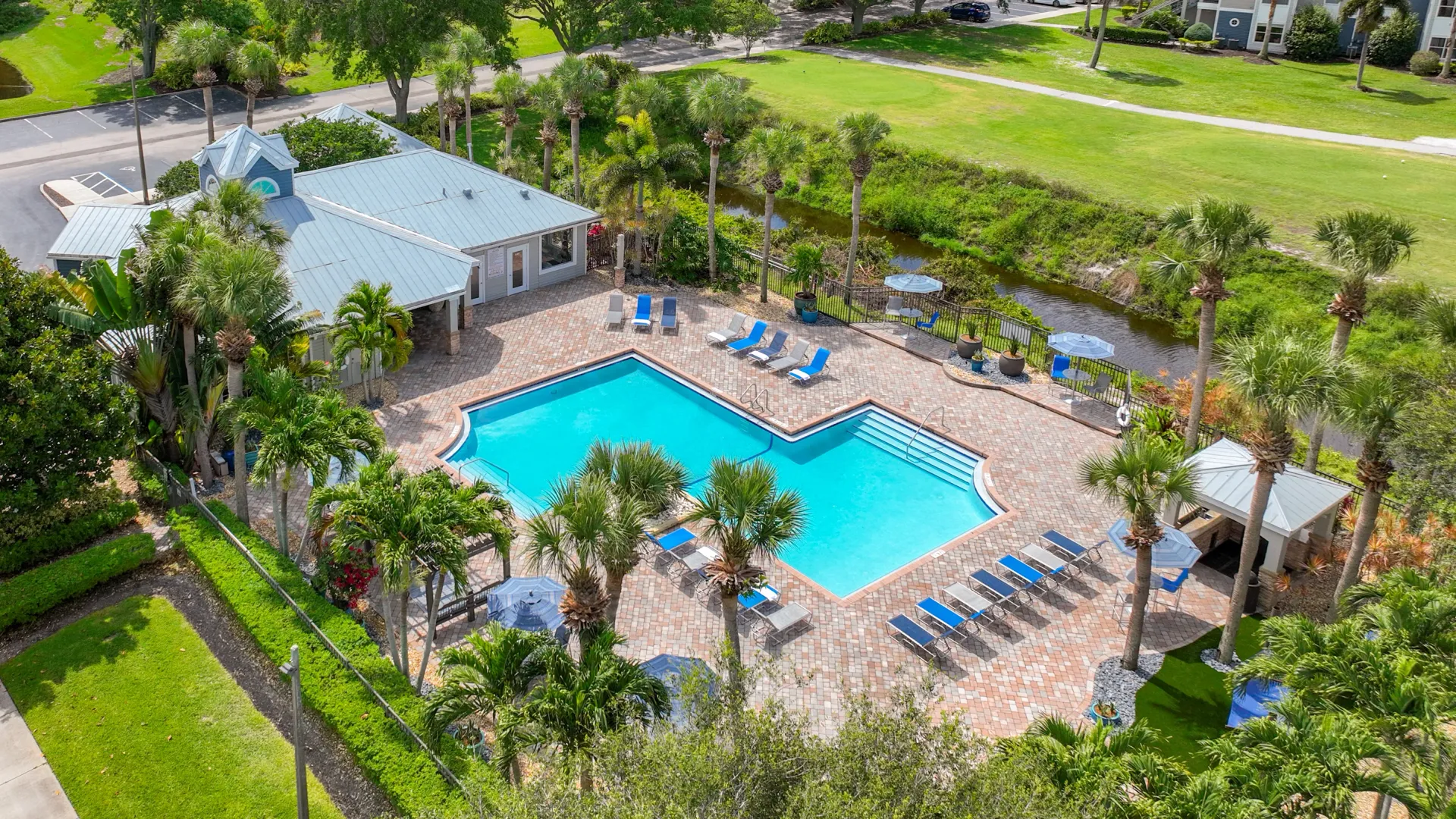 Aerial view of the resort-style pool at Beachway Links Apartments featuring an expansive sundeck, lounge chairs, palm trees, and landscaped grounds.