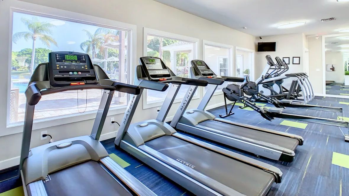 A wall of cardio machines – including multiple treadmills, ellipticals, and row machines - facing the pool deck, seen through massive picture windows, offers residents a captivating view while working out.