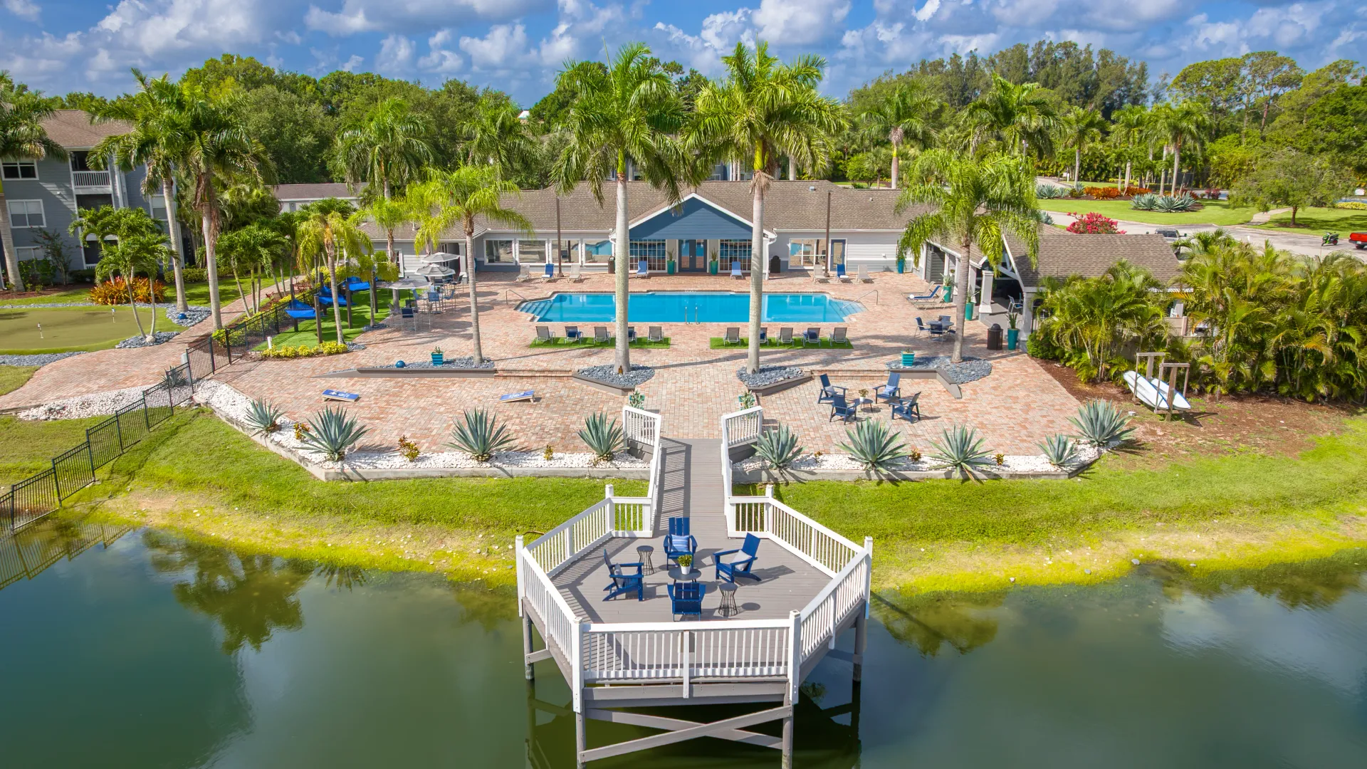 Aerial view of Grand Oaks Apartments’ resort-style pool area with palm trees, lounge chairs, and a lakeside deck with seating.
