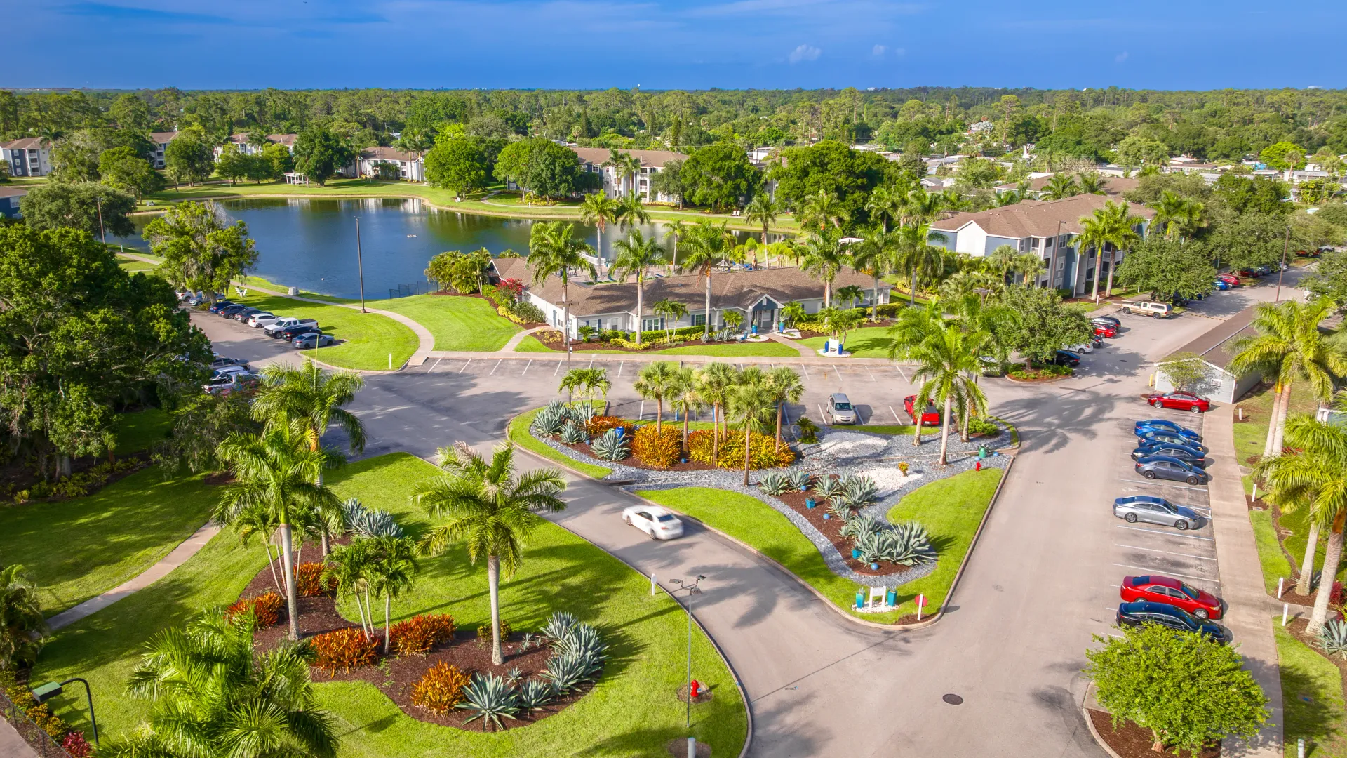 Aerial view of Grand Oaks Apartments entrance with landscaped roundabout, palm trees, and community buildings near a lake.