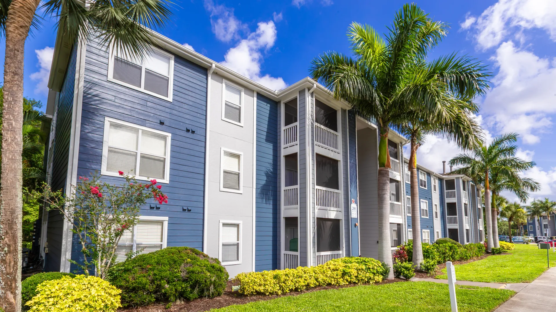 Three-story apartment building at Grand Oaks Apartments with screened balconies, palm trees, and landscaped grounds.