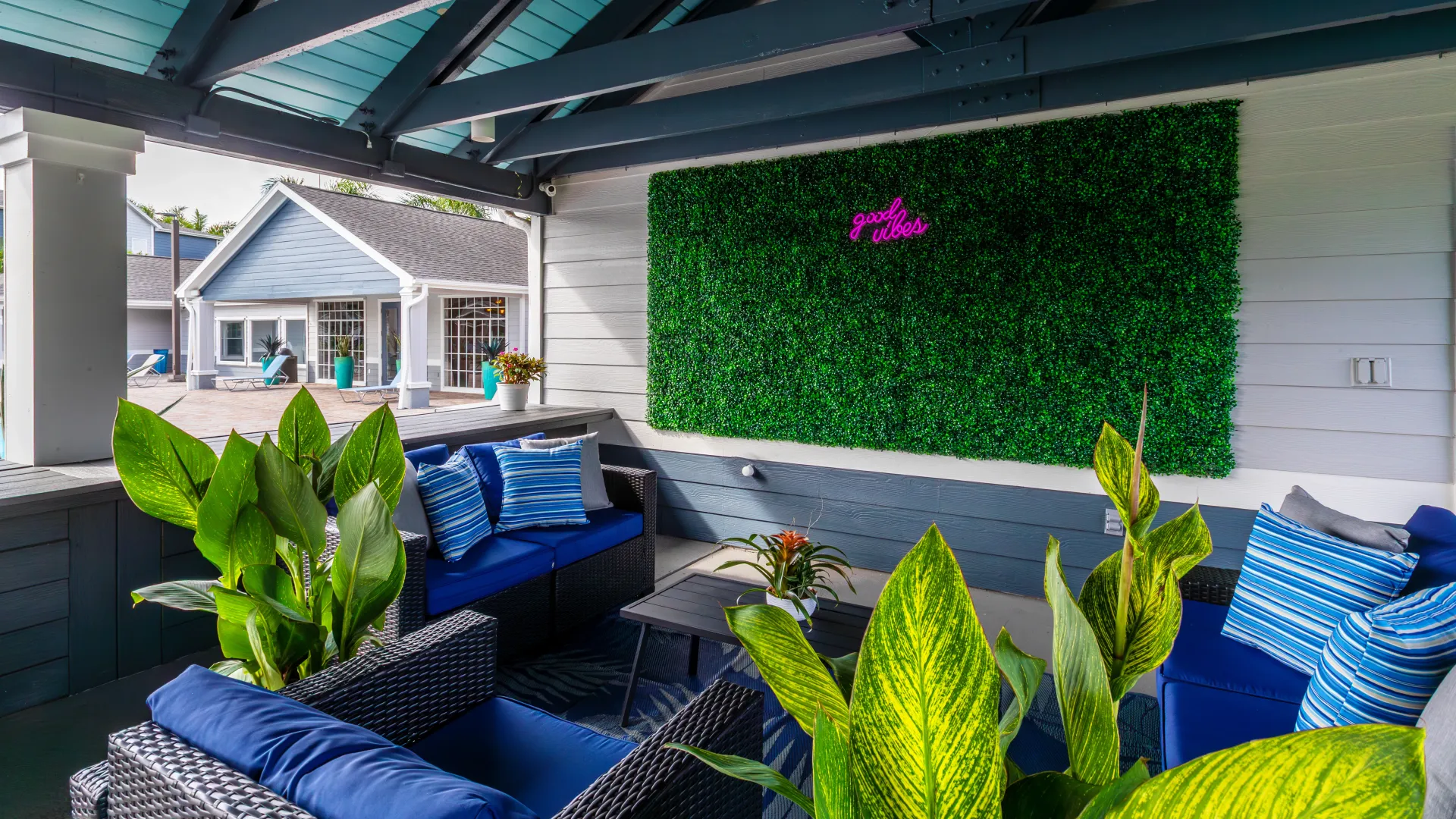 Covered outdoor lounge at Grand Oaks Apartments with wicker seating, tropical plants, and a “good vibes” neon sign.