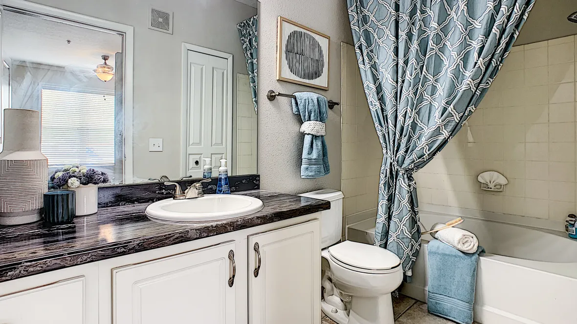 A bathroom with upgraded white cabinetry and black fusion countertops, extended mirror with vanity lighting, and garden tub.