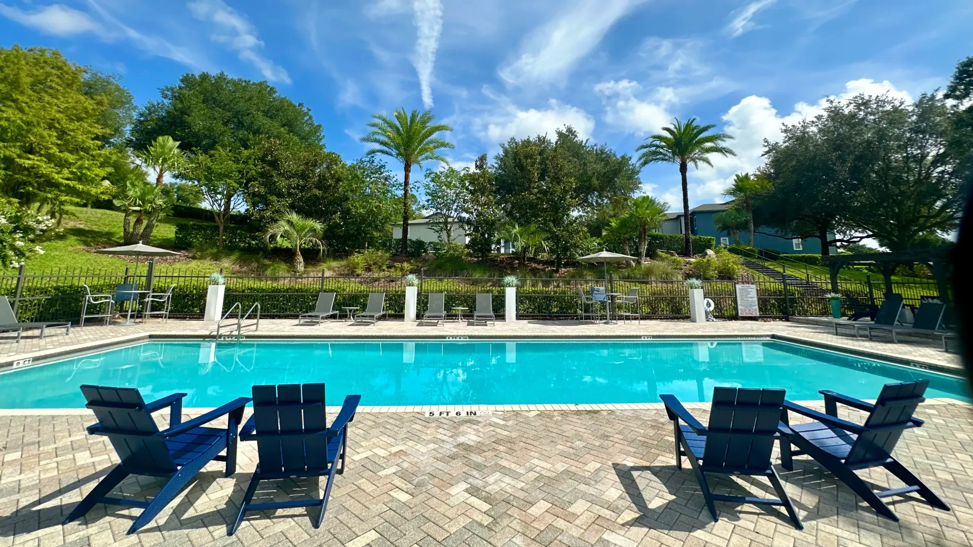 Poolside area featuring blue lounge chairs, surrounded by lush greenery and palm trees under a clear blue sky.