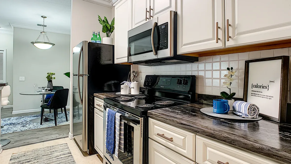 A view of the side of the kitchen that features a stainless steel stove, microwave, and refrigerator, with white cabinetry and black fusion countertops, and white subway tile backsplash.