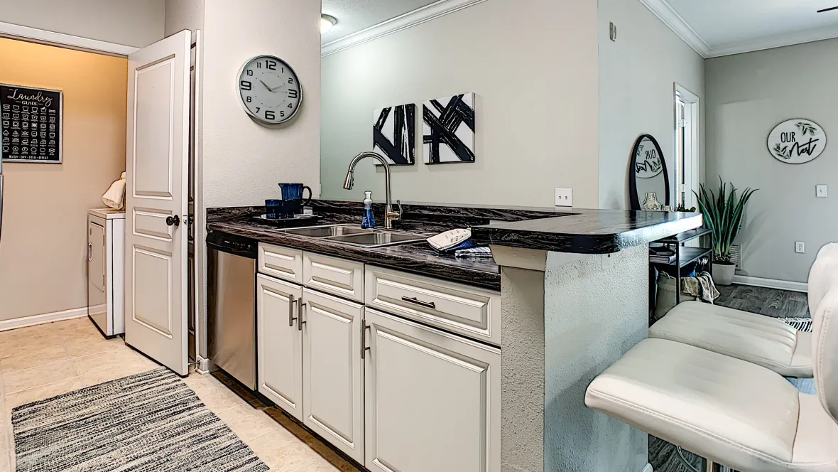 A view of the side of the kitchen that features a stainless steel dishwasher, large sink, expansive breakfast bar, which opens into the living room space.