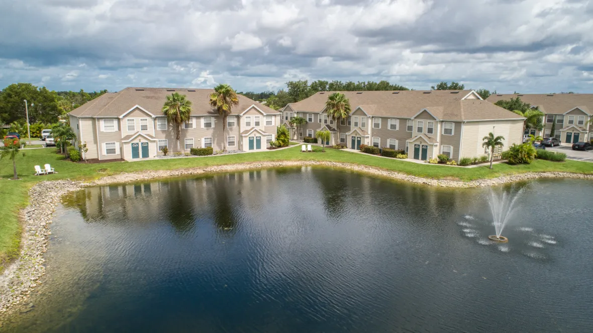 A serene aerial view showcases the glistening lake and a tranquil fountain next to well-maintained apartment buildings, with plenty of lush greenspace to enjoy.