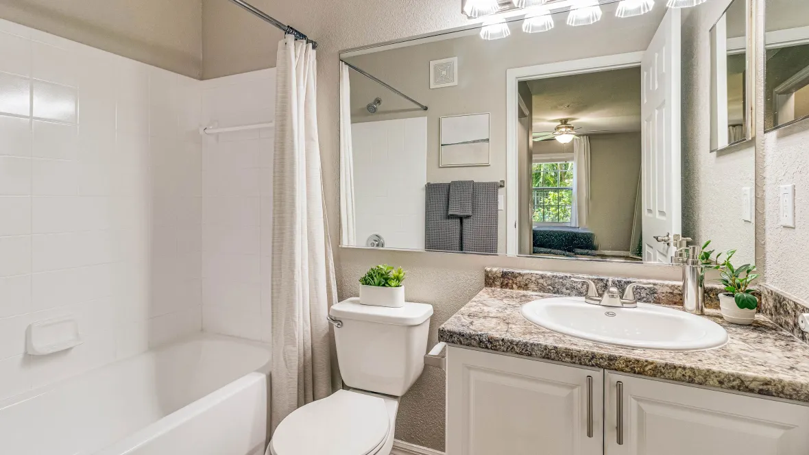 A bathroom featuring a large mirror reflecting radiant lighting from contemporary overhead fixtures and natural light from the adjacent bedroom window.