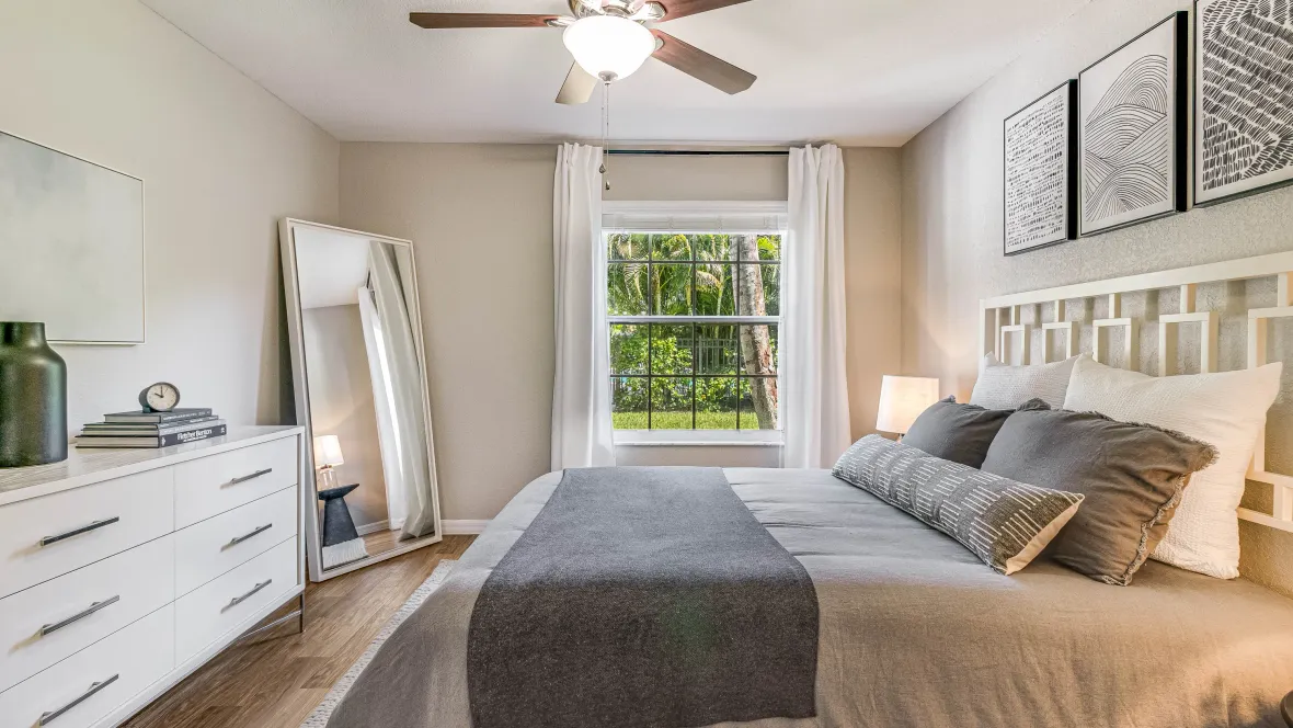 A serene bedroom, aglow with natural light from a large picture window, slick wood-like floors, and an illuminated ceiling fan for an elegant ambiance exuding comfort.