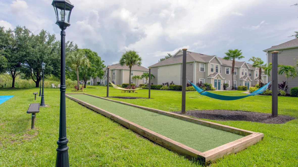 A delightful area within a grassy field set up for leisure activities including two hammocks, a bocce ball court, a shuffleboard court, and park benches with three dedicated light poles to enjoy activities in the evening hours.