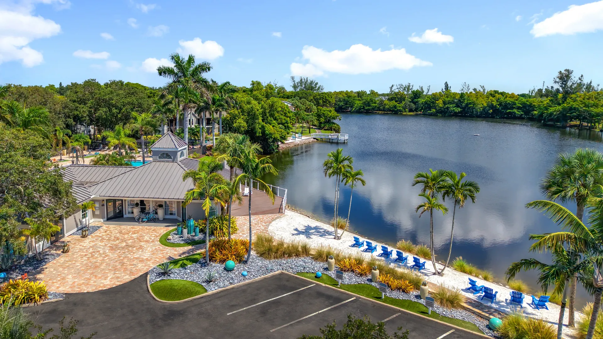 Aerial view of River Reach's lakeside clubhouse with a sandy beach area, palm trees, and blue Adirondack chairs along the lakefront.