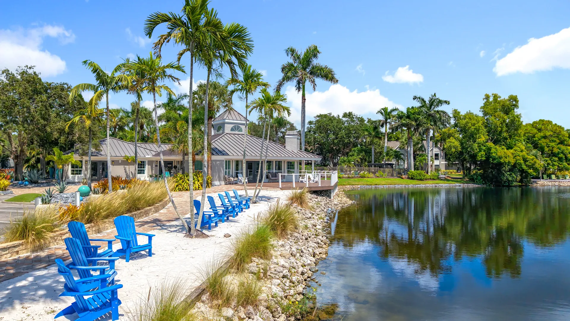 Lakeside view of River Reach's private white sand beach with blue Adirondack chairs, palm trees, and the clubhouse along the shoreline.