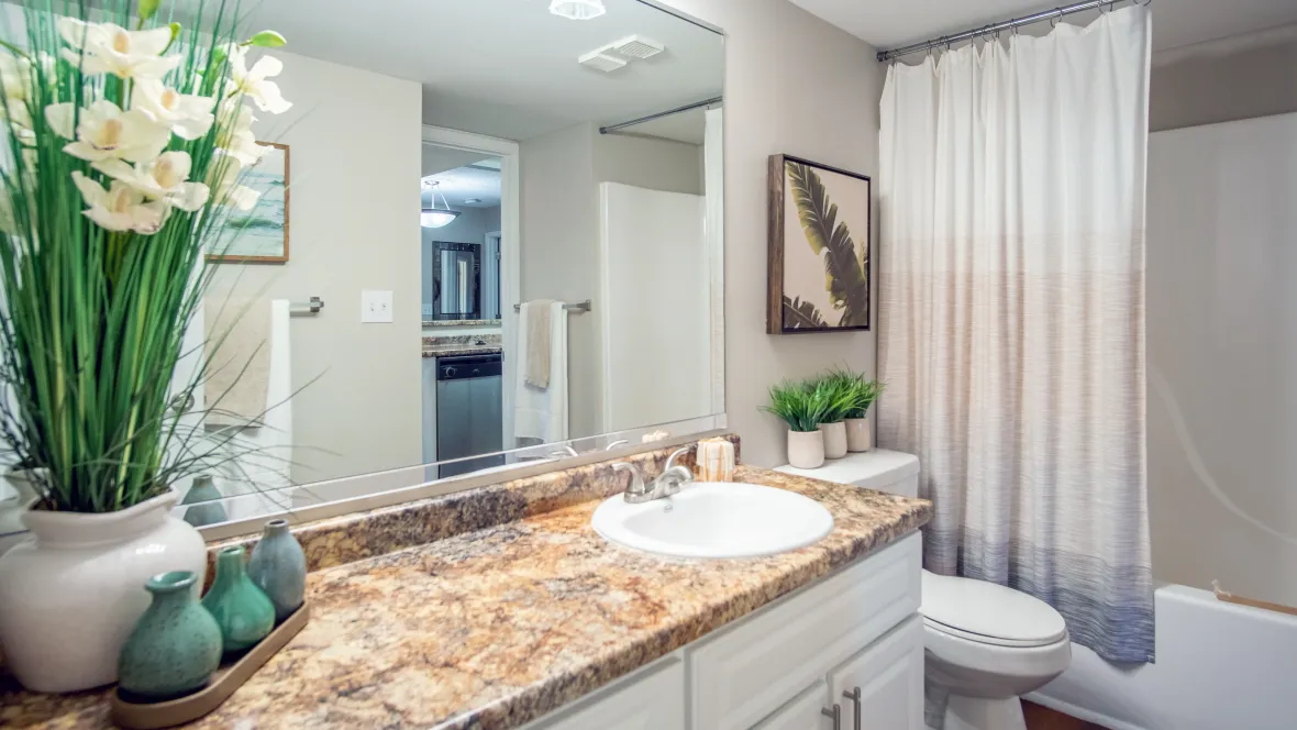 A restroom featuring white shaker cabinets and drawers below a sprawling countertop left of the sink, with an extended mirror and bright overhead vanity lighting, next to a white tile surrounded shower/tub combination.