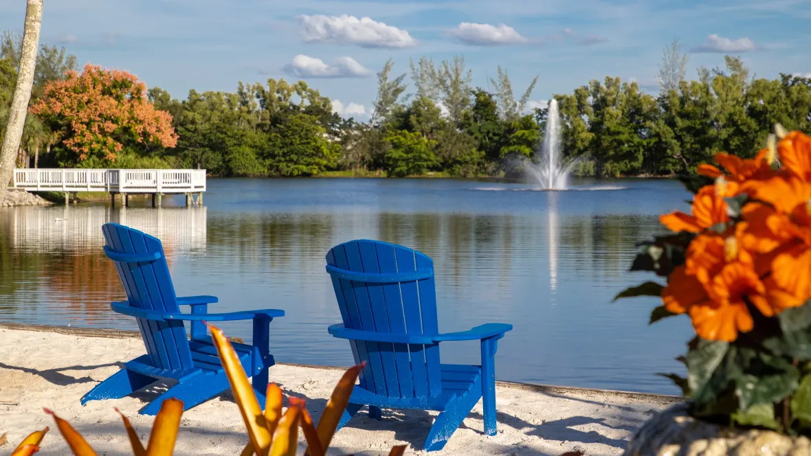 Two vibrant blue Adirondack chairs positioned in the sandy beach area facing the soothing lake fountain with the tree lined lake and peaceful blue skies creating a scene of serenity.