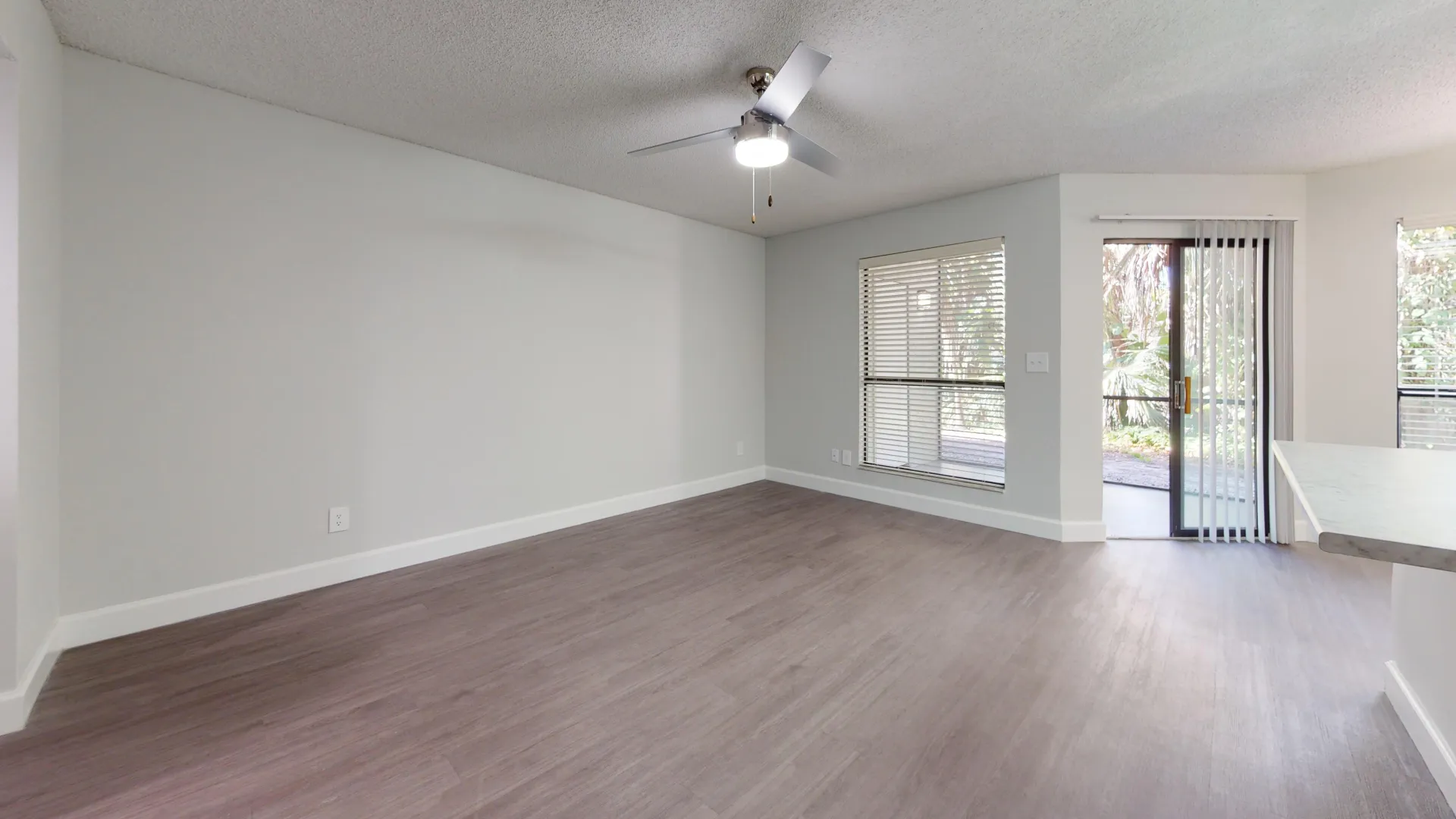 Spacious living room in the Martinique floor plan with natural light coming through large windows and sliding glass doors, leading to a private patio. The room has modern flooring and a ceiling fan.