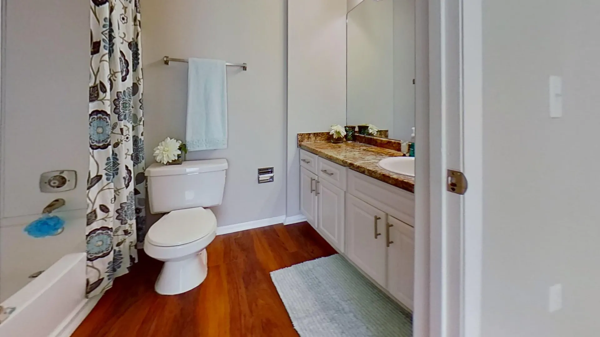 Bathroom in the Martinique Autograph floor plan, showcasing a granite countertop, modern cabinetry, and hardwood-style flooring with a shower curtain and decorative towel.