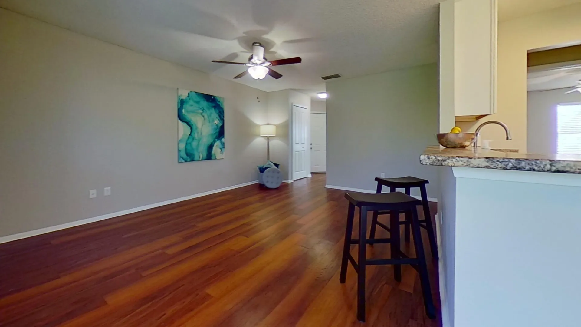 A spacious living room in the Martinique Autograph floor plan, featuring wood-style flooring, a ceiling fan, modern decor with a blue abstract painting, and bar seating at the kitchen counter.