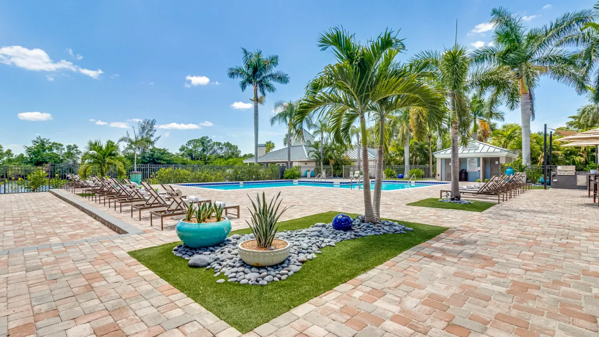 An expansive, paved sundeck with cascading palm trees and pockets of astroturf and intriguing landscape design, offering residents a serene spot to relax and soak up the sun in a vibrant setting. 