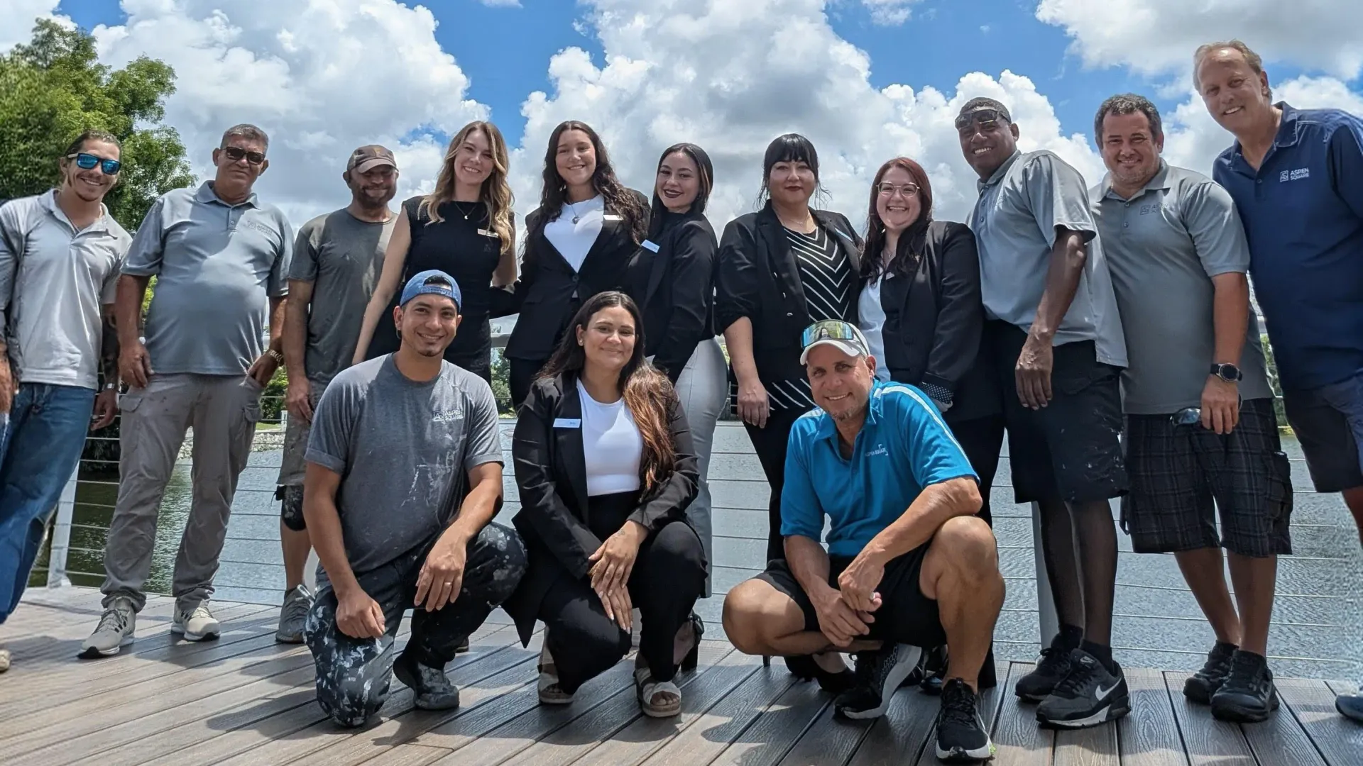 Group photo of the River Reach Apartments team, smiling outdoors on a sunny day, wearing branded uniforms and casual attire.