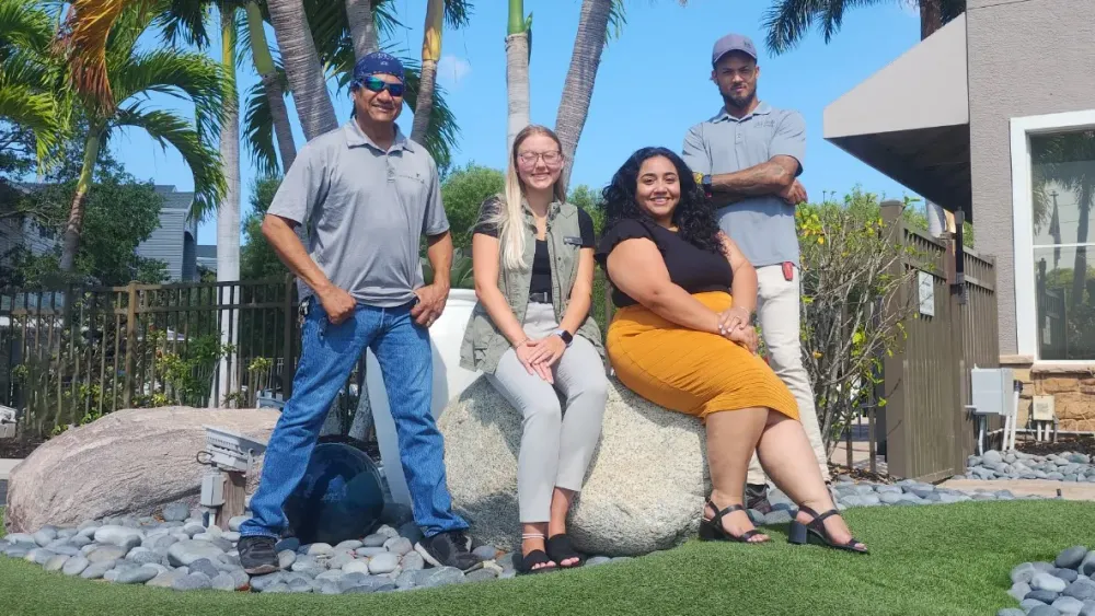 Four team members at Somerset Palms Apartments smiling outside near the clubhouse, standing and sitting on decorative boulders surrounded by palm trees and landscaping.