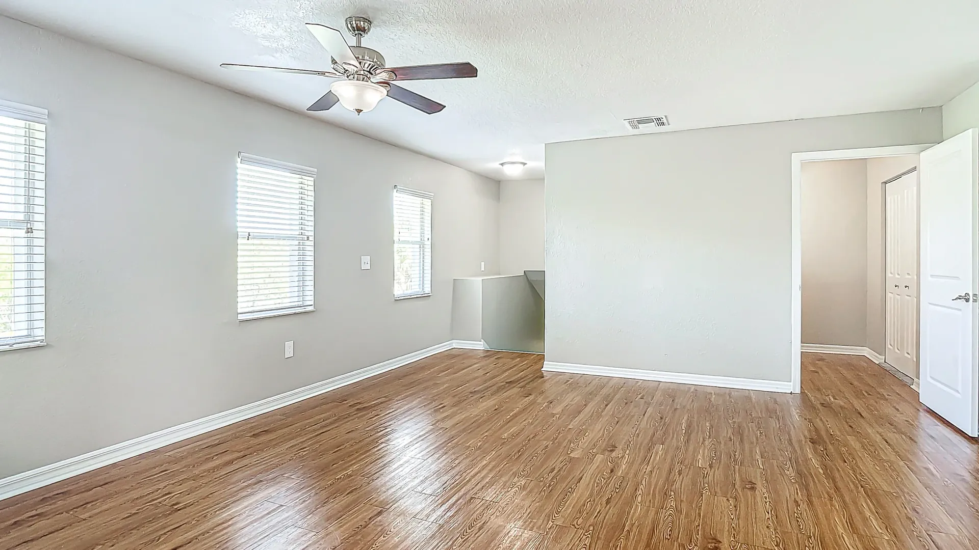 A well-lit living area with wood-style flooring, multiple windows with blinds, a ceiling fan, and neutral-colored walls, offering a versatile and inviting space.