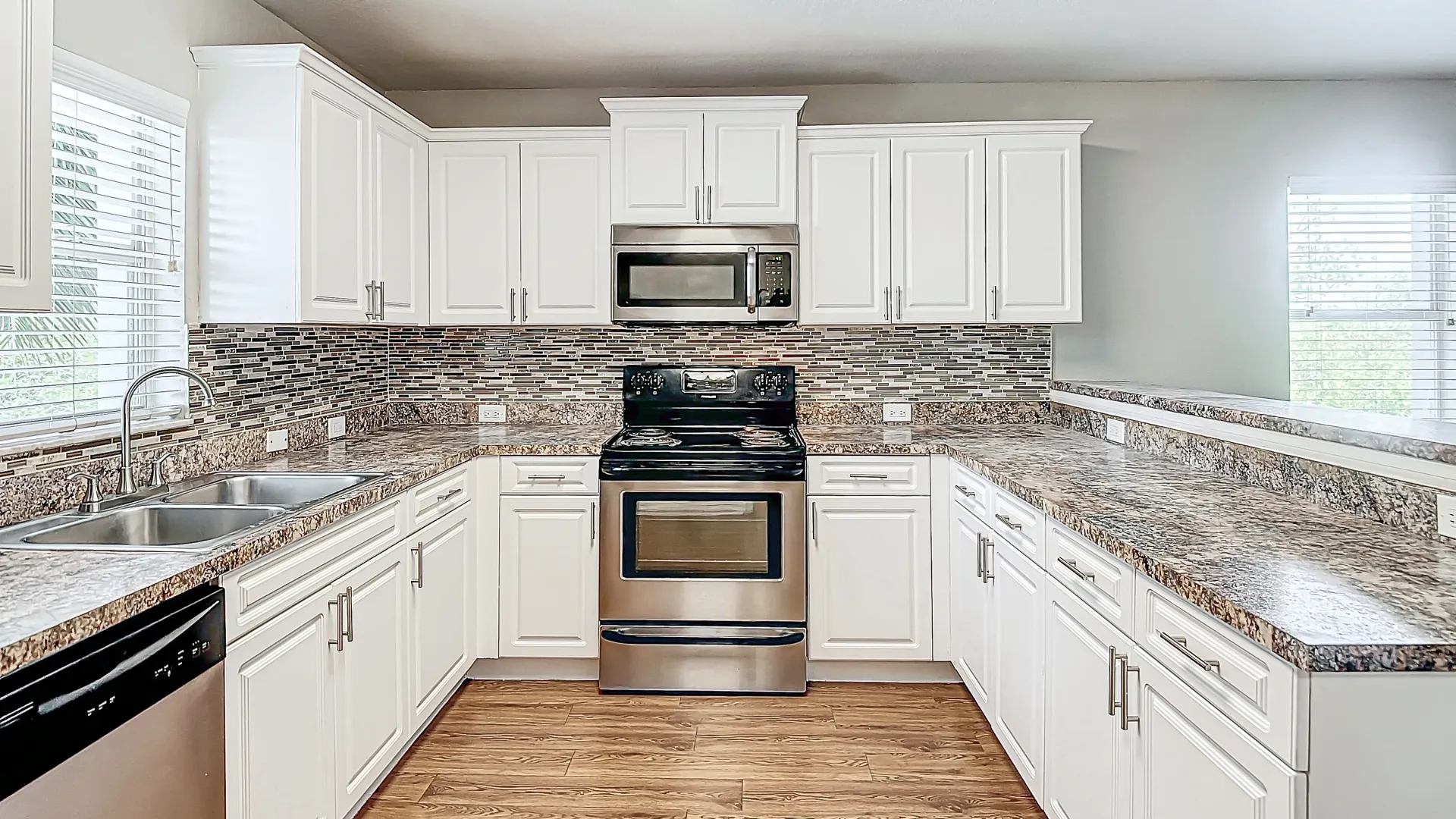 A bright, modern kitchen with stainless steel appliances, white cabinetry, granite countertops, and a sleek mosaic tile backsplash.