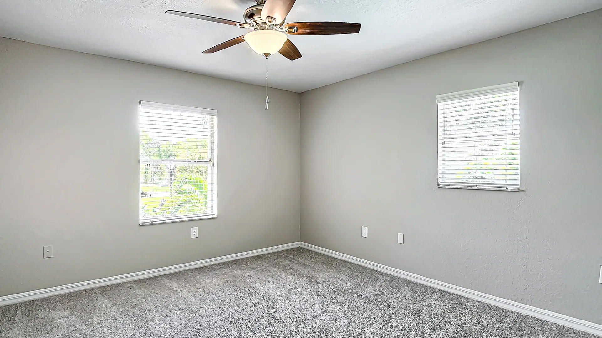 A well-lit bedroom with soft gray carpeting, neutral-colored walls, and large windows with blinds, allowing plenty of natural light to fill the room.