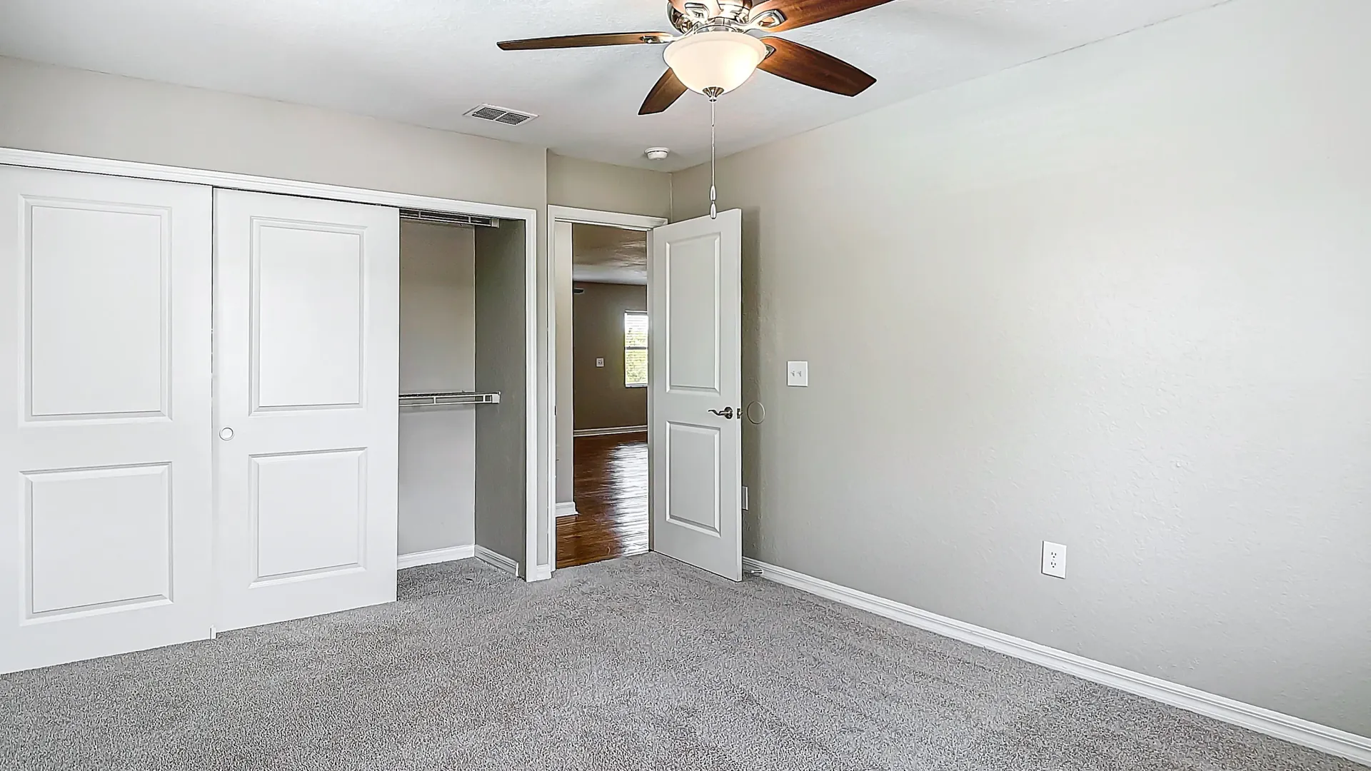 A bright bedroom with gray carpet, a ceiling fan, and a large closet with sliding doors, providing ample storage space.