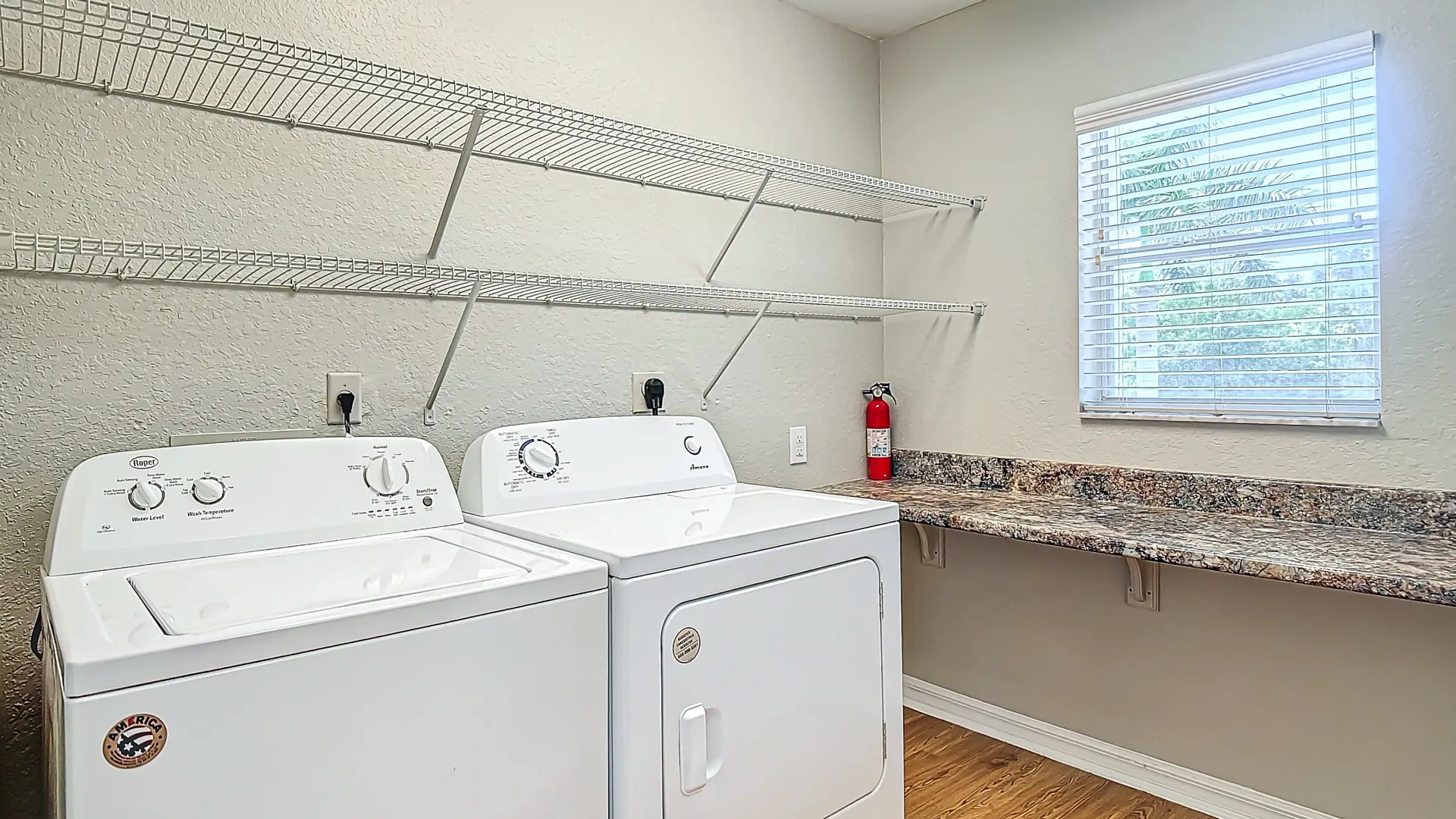 A laundry room with a washer, dryer, built-in wire shelving, and a large countertop under a window, located in an apartment.