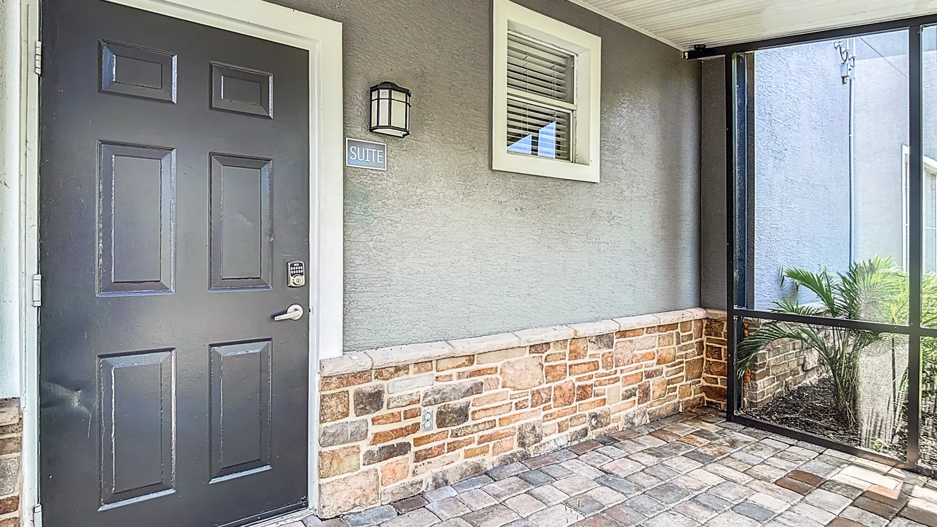 A close-up view of a The Suite floor plan entrance, featuring a dark-colored door, stone accents on the lower wall, and a screened patio area.