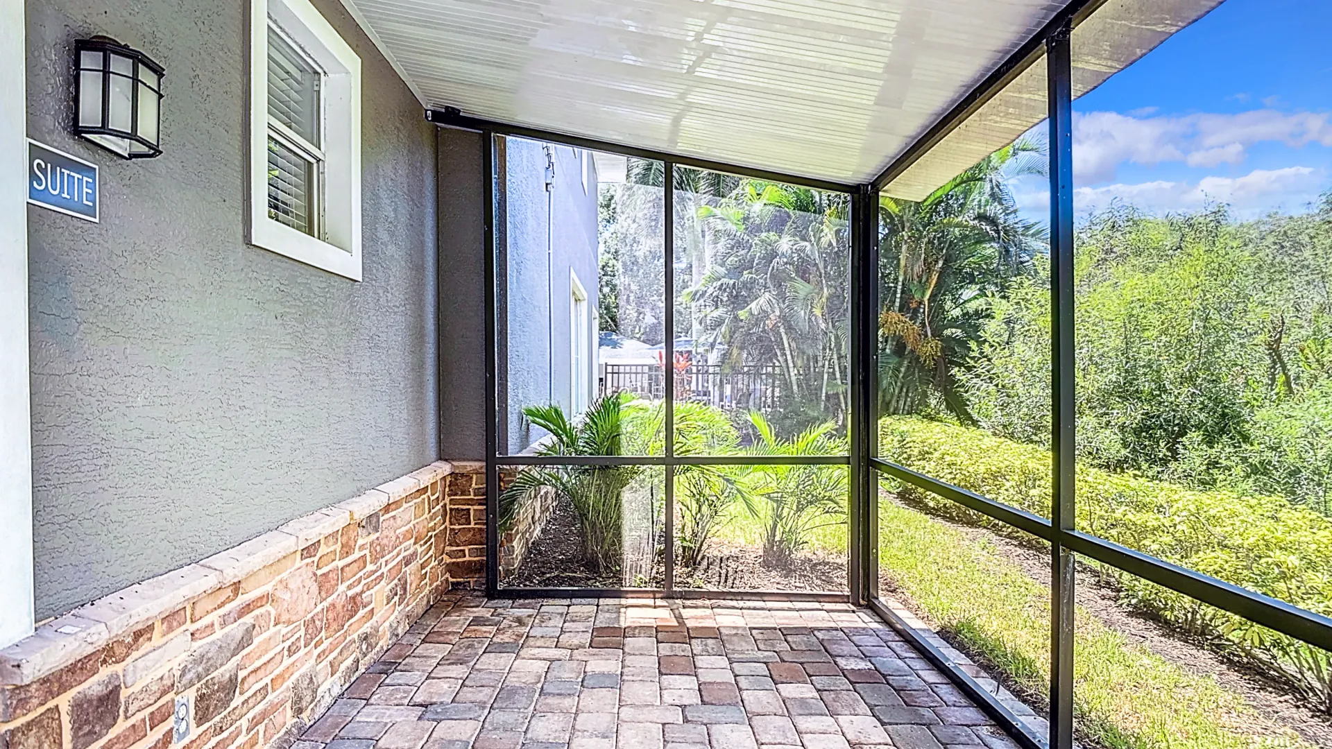 A screened patio featuring stone accents on the lower wall, paver flooring, and a view of lush greenery outside. The area is shaded, providing a relaxing outdoor space.