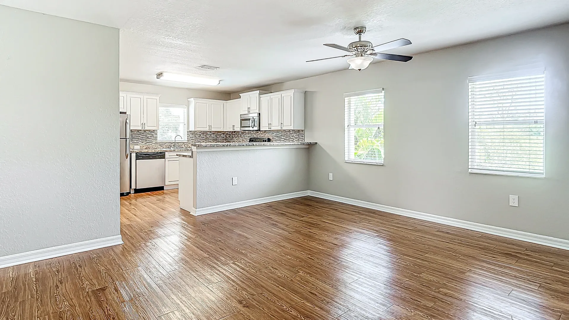 An open-concept living area with hardwood floors and large windows, connecting to a modern kitchen with stainless steel appliances and white cabinets.