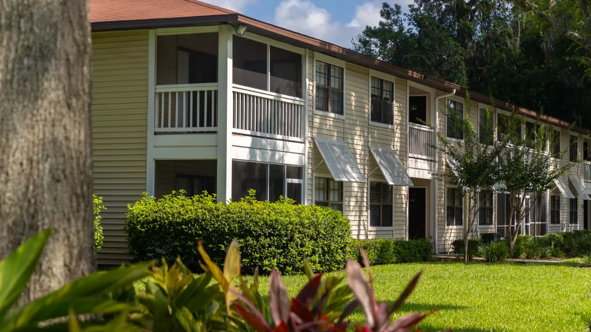 Exterior view of two-story apartment buildings, encircled by lush greenery, shrubbery, and towering pine trees, creating a peaceful and heartwarming atmosphere.