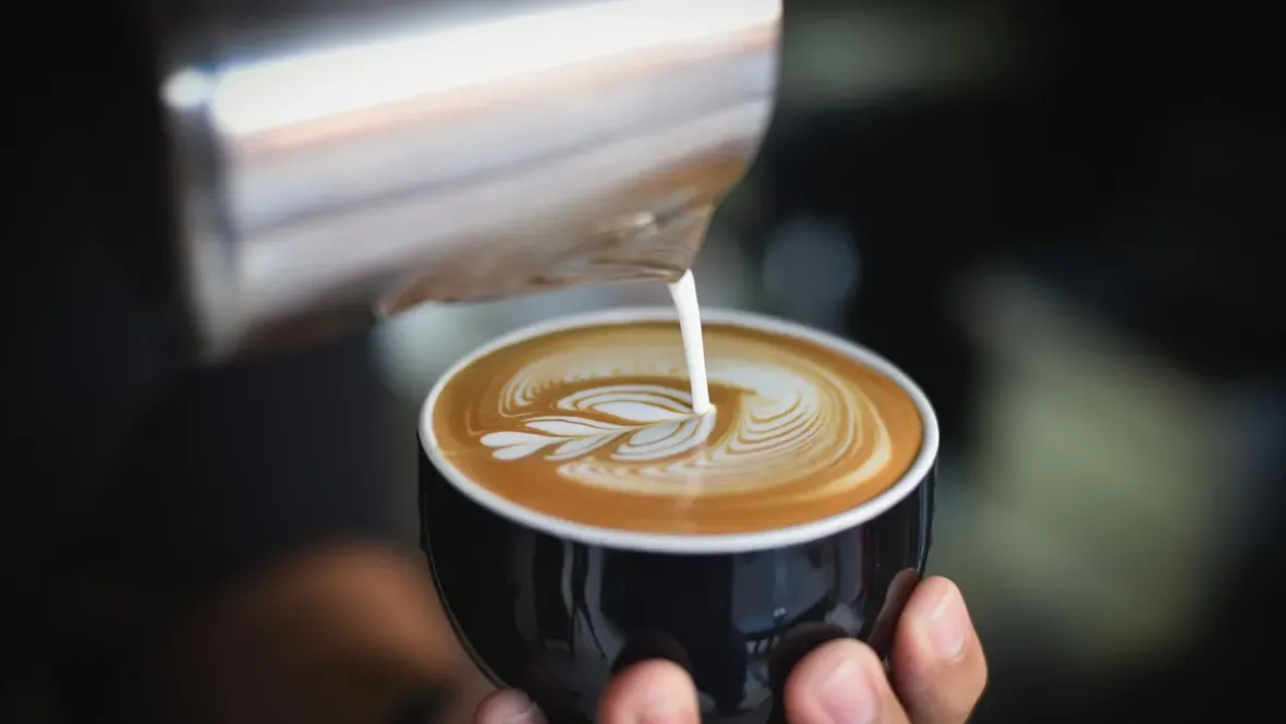 A barista pouring steamed milk over a cappuccino in a black mug.