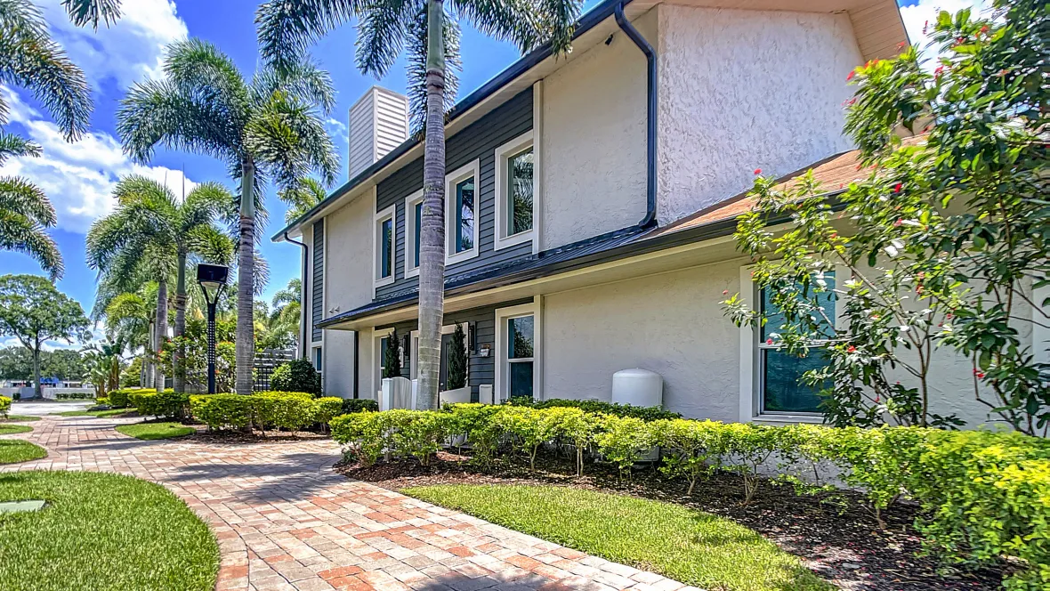 The exterior of an apartment building, lined with plush green grass and palm trees.