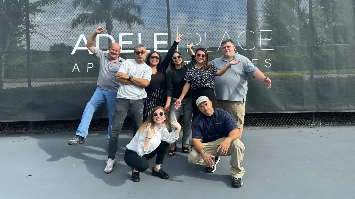 The Adele Place team posing in the tennis court with a banner behind them that reads "Adele Place".