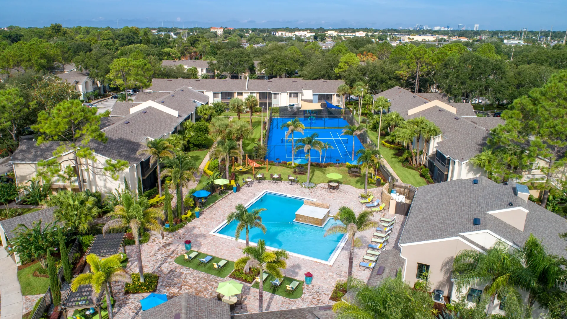 An aerial view of our splendid community, nestled among lush trees, highlighting the expansive sundeck and vibrant tennis court that mirrors the sparkling blue pool.