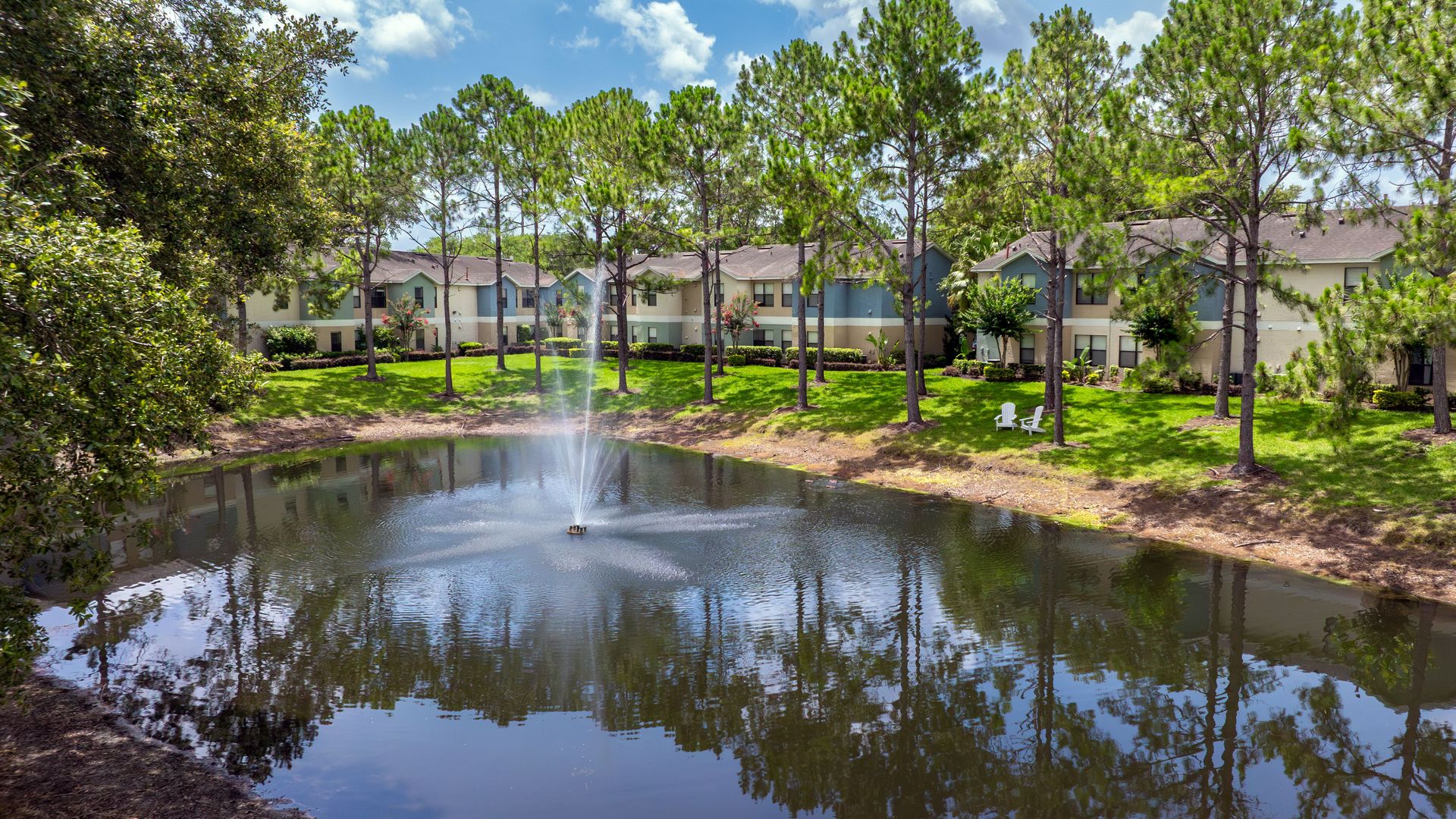 Serene pond with water fountain surrounded by pine trees and apartment buildings at Harper Grand.