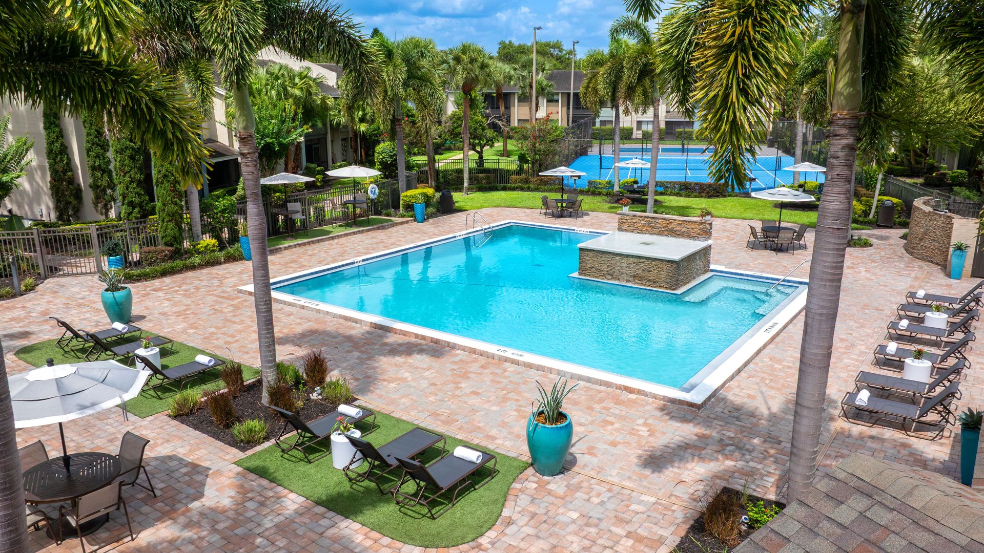 View of Harper Grand’s pool with lounge chairs, shaded tables, palm trees, and a raised water feature.