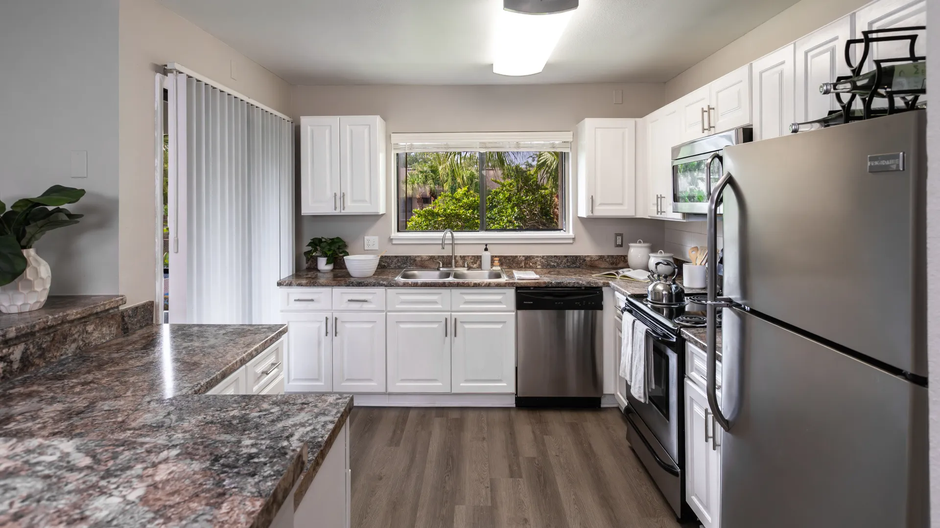 Modern apartment kitchen with stainless-steel appliances, white cabinets, large window, and wood-style flooring.
