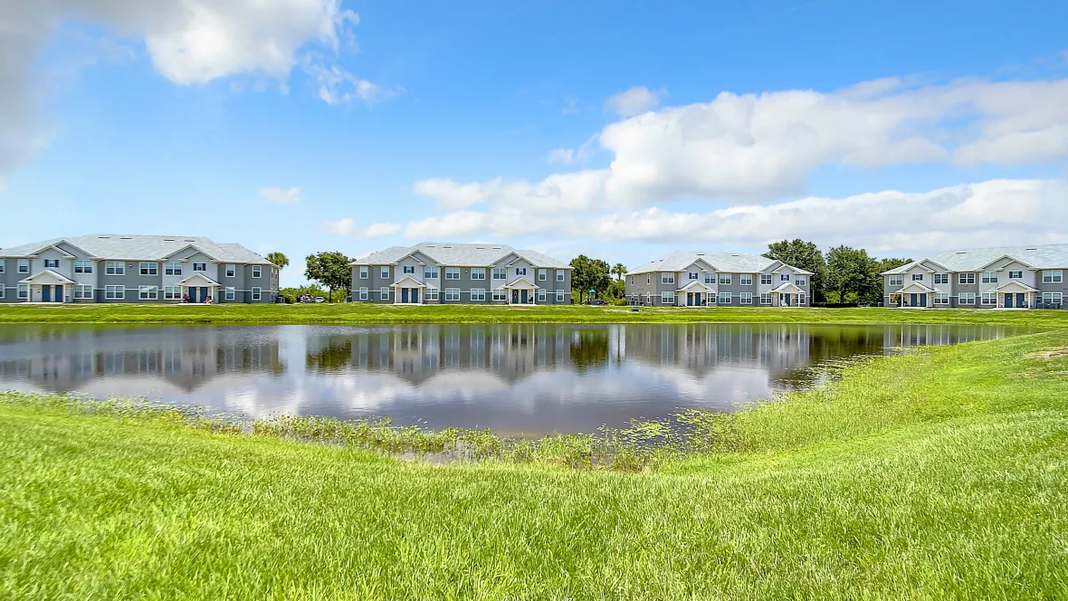 A lake surrounded by lush, green grass with apartment buildings visible across the other side of the water.