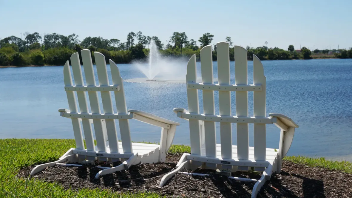 Two white Adirondack chairs facing the glistening lake and trees in the background, offering a restful space for residents to unwind.