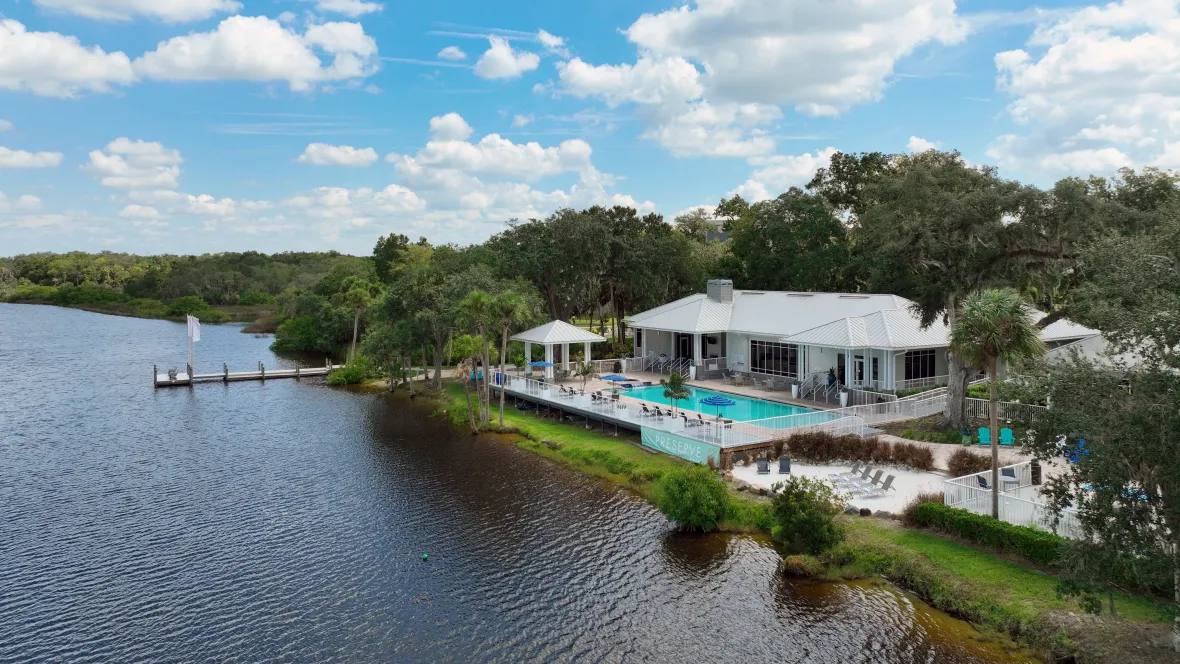 A captivating aerial view over the river showcasing the community’s delightful beach area, stunning pool, extended riverside deck and fishing pier offering a blend of natural beauty and lavish amenities.