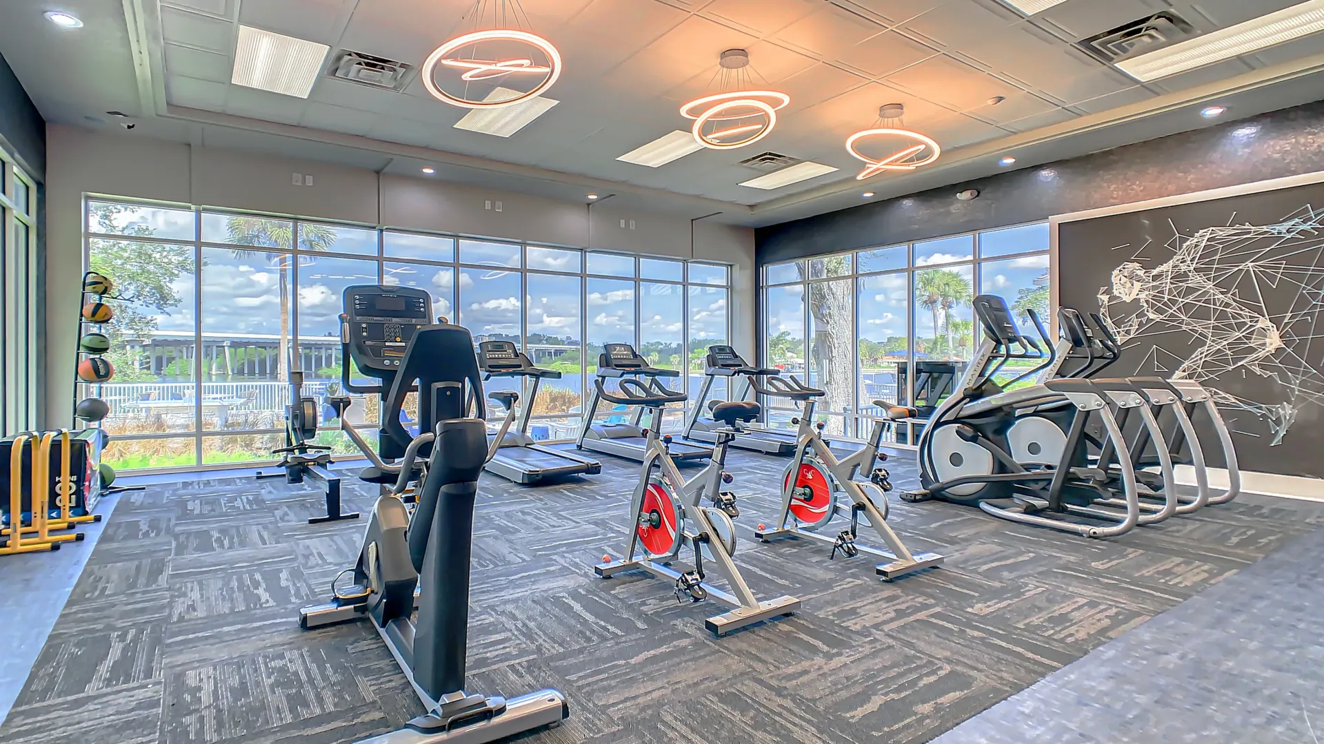 A jaw-dropping gym with contemporary lighting, wall-to-wall windows showcasing panoramic views of the pool and river, and rows of equipment including ellipticals, treadmills, rowers, and spinning bikes.