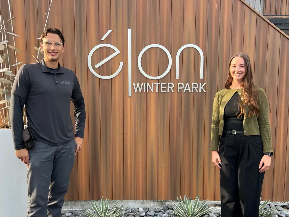 Two Elon Winter Park team members standing outdoors beside the community’s branded Elon Winter Park wall.