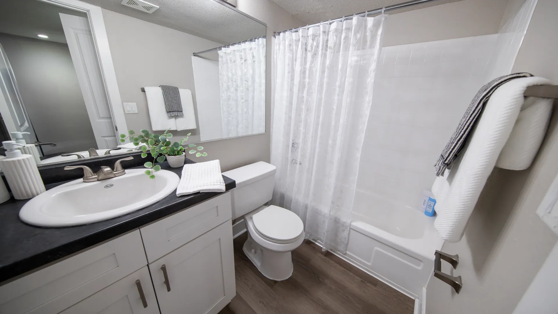 A modern bathroom at Elon Winter Park Apartments featuring a white sink, toilet, and a shower with a white curtain, accented by a black countertop and wood-style flooring.