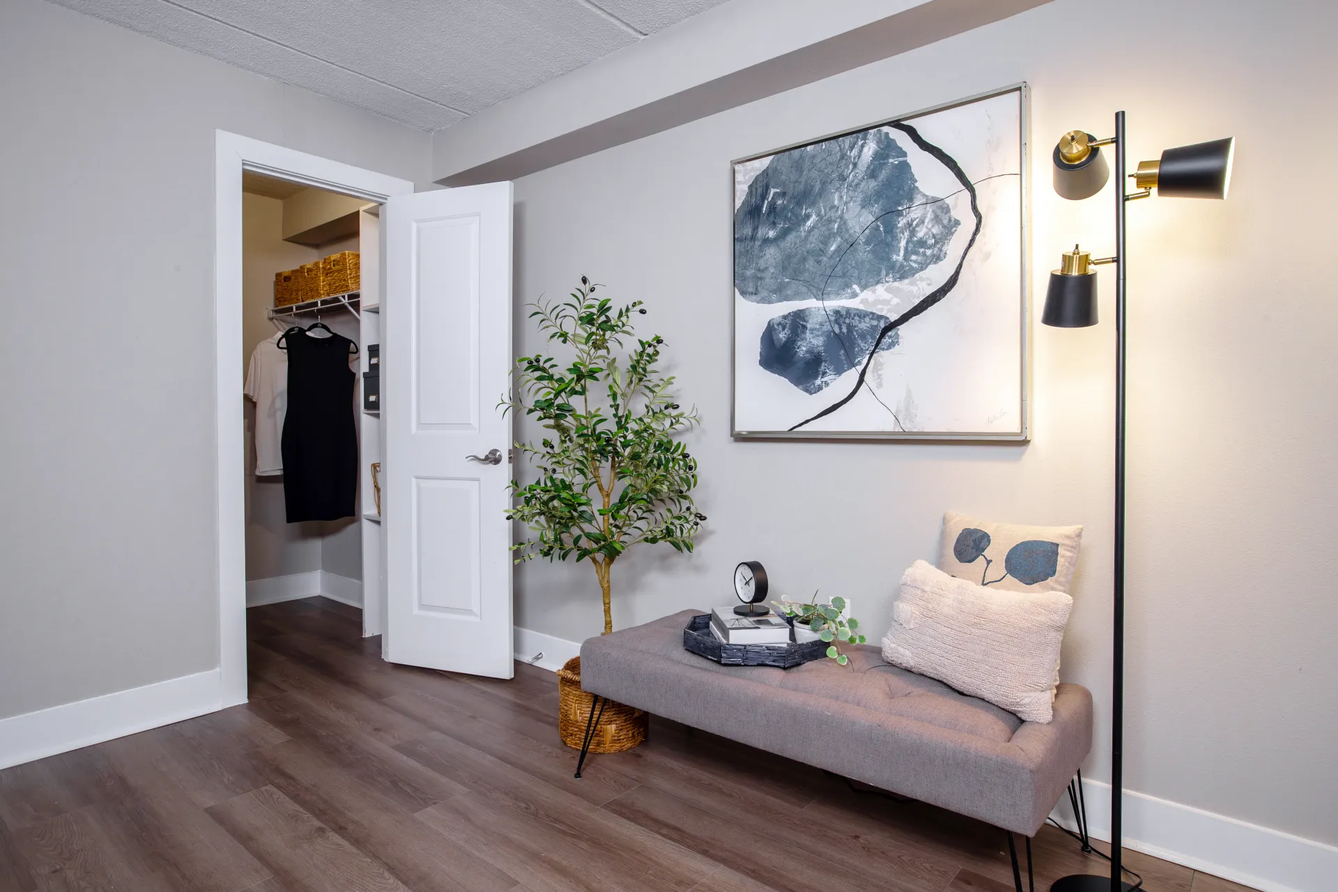 A bedroom with modern wood-style flooring, a cushioned bench, a tall lamp, decorative artwork, and an open walk-in closet in the background.