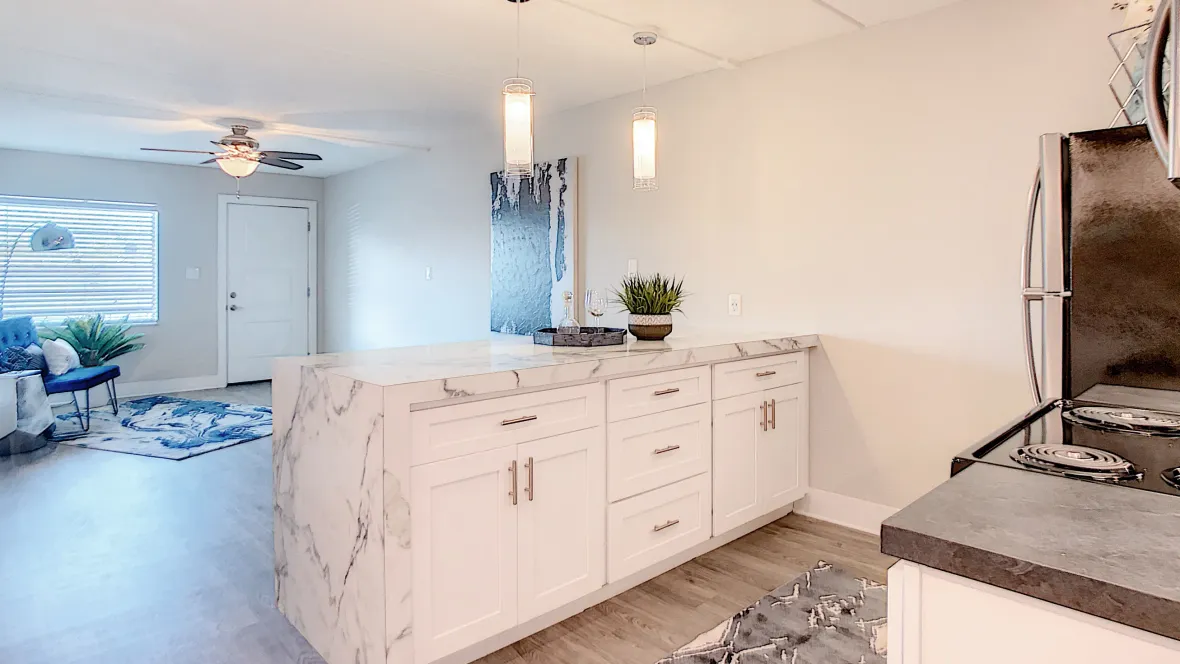 A full view of the breakfast bar, lined with Carrara marble inspired countertop and featuring additional upgraded, white cabinetry underneath, with the living room space visible in the background.