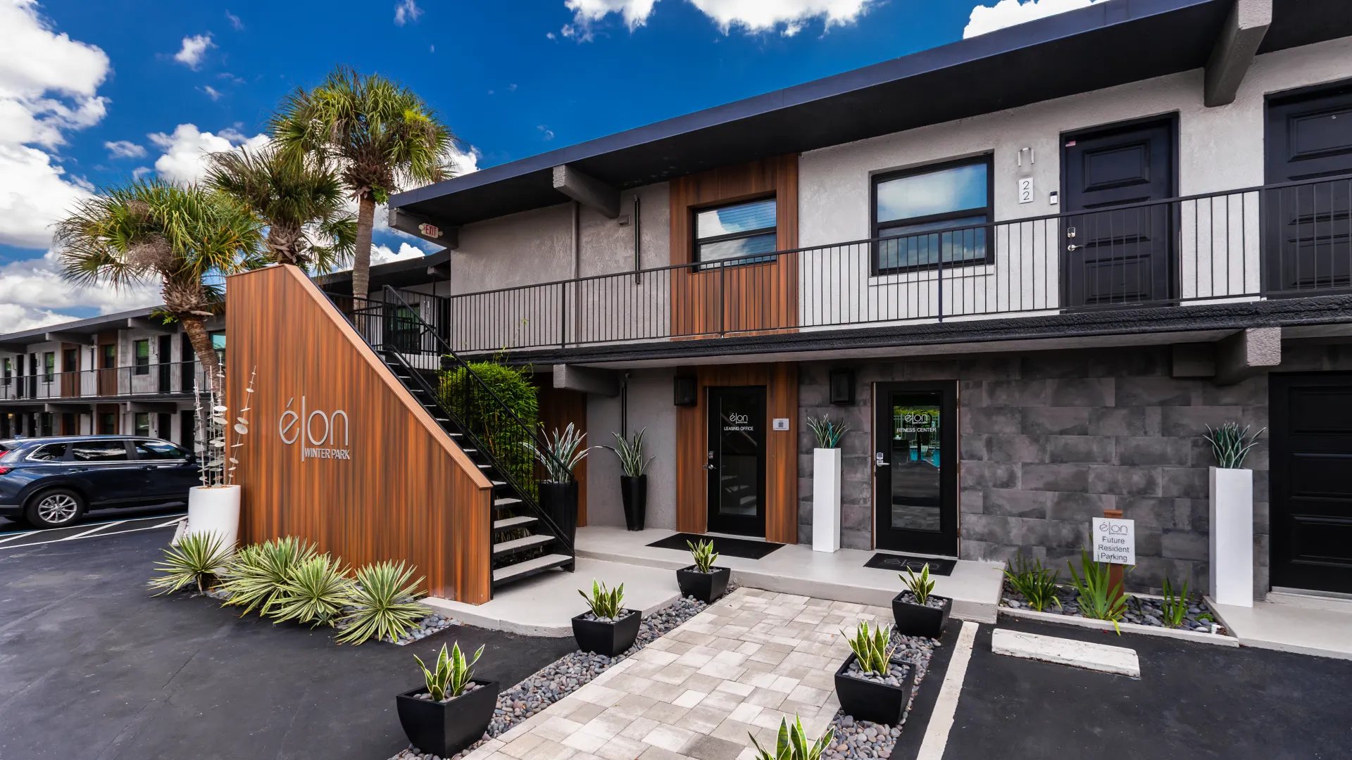 Exterior of Elon Winter Park Apartments featuring modern architecture, landscaped walkways, and signage for the leasing office and fitness center.