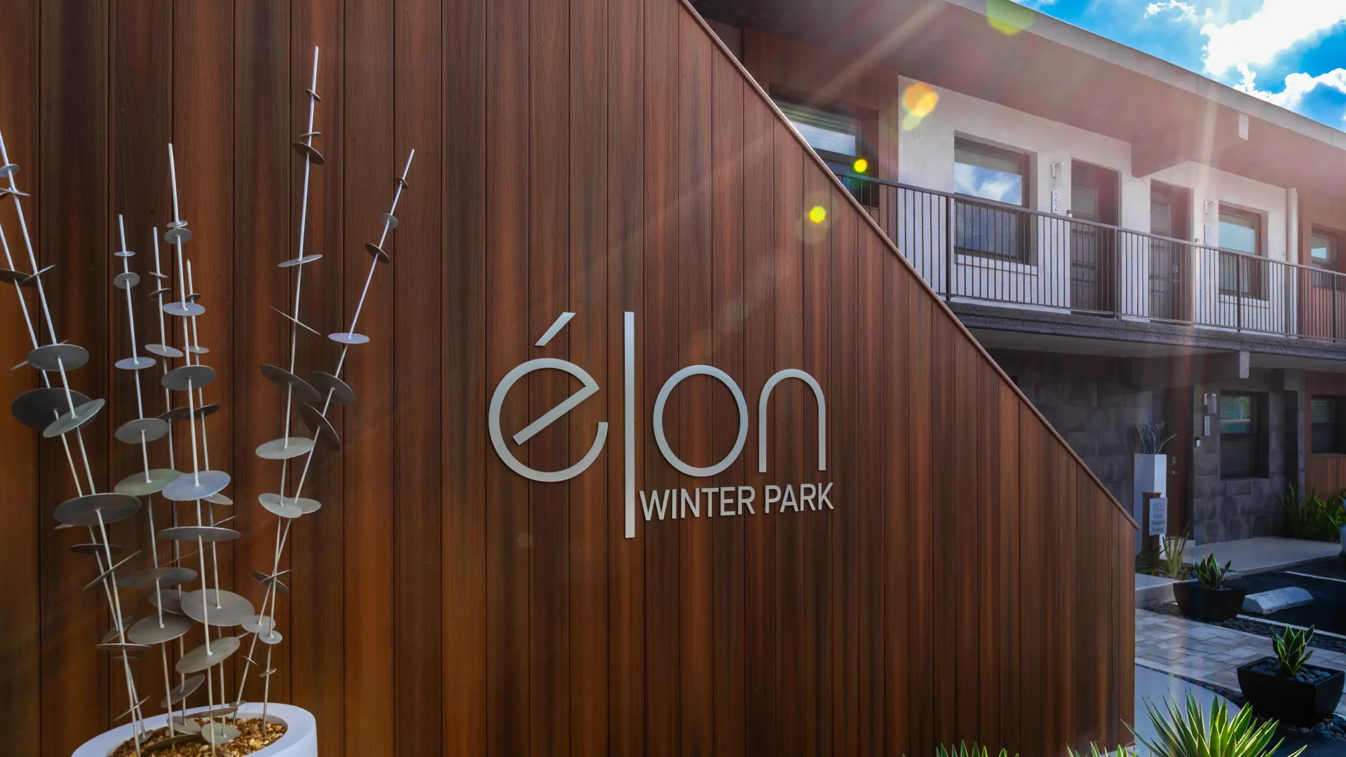 Close-up of the Elon Winter Park Apartments signage with modern architectural design and artistic metal sculptures, set against a wood-paneled exterior.